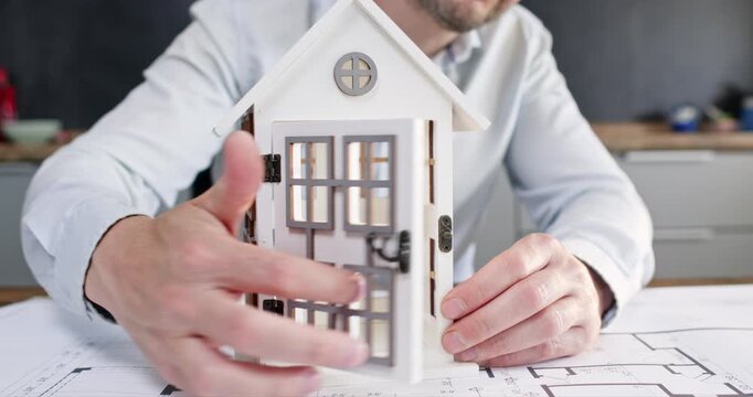 Man realtor in white shirt sits at table and turns miniature model house. Male architect carefully opens miniature front door and explains layout of actual home
