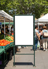 A blank vertical sign mockup on a green stand at a busy outdoor farmers market with shoppers.