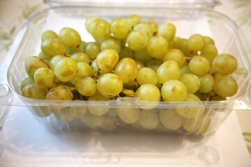 Fresh green grape bunch in a transparent plastic container, photographed close-up in a market or home setting.