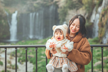 Happy Asian mother holding excited little daughter enjoying trip near Shiraito waterfall in Japan...