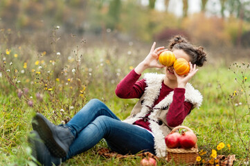 Playful Smiling girl sitting in grassy autumn field holding small pumpkins over her eyes, with a basket of apples beside her, celebrating Thanksgiving outdoors Autumn Harvest.