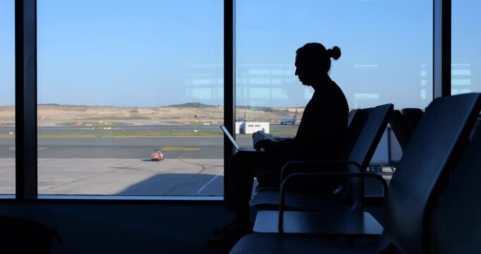 Man turns on his laptop and checks webcam to join work meeting. He sits in waiting area at large airport, silhouetted shot against window. Concept of modern lifestyle with popularity of telecommuting