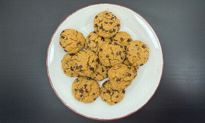 Homemade chocolate chip cookies served on white ceramic plate with brown rim, arranged on dark wooden table for dessert, snack or sweet treat presentation