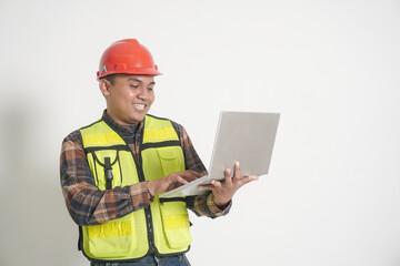 Asian construction worker wearing safety vest and helmet, smiling cheerfully while holding a laptop, representing modern technology in construction. Isolated image on white background
