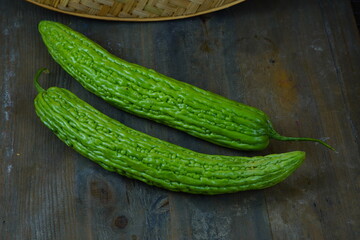 Two vibrant green bitter melons are placed on a rustic wooden table, with a woven basket visible in...