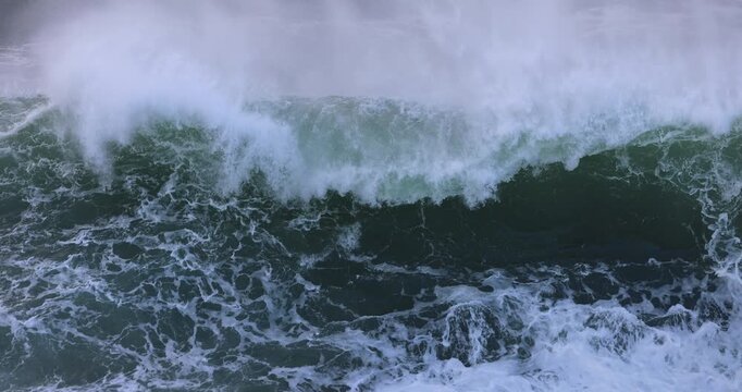 An large ocean storm wave curls and breaks over a coastal rock ledge, taking the appearance of a huge waterfall, before exploding skyward into a giant wall of white water. Captured in slow motion.