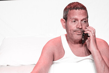 Adult male making a funny gesture while talking on phone on bed on white background