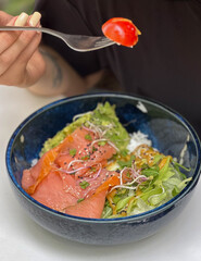 A woman is eating a bowl with red fish, rice and mango, holding a fork with a tomato. Outdoor setting in natural daylight, healthy lunch or brunch in a modern lifestyle context.


