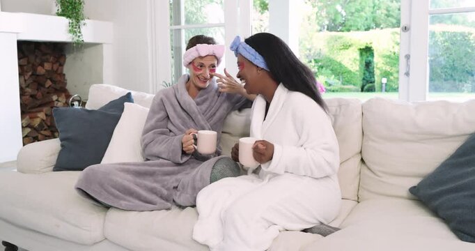Diverse female friends in robes settling on sofa, sipping mugs comparing eye gel patches for spa