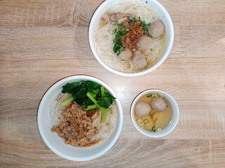 A top-down view of two bowls of bakso and one bowl of noodles, with meatballs, minced chicken, and vegetables on a wooden table