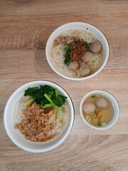 A top-down view of two bowls of bakso and one bowl of noodles, with meatballs, minced chicken, and vegetables on a wooden table