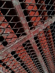 A close-up, angled view of a diamond-shaped metal mesh and a red painted support beam, with a red brick background