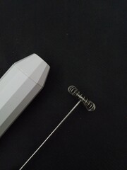 A simple, minimalist flat lay of a battery-operated milk frother with its two different whisk attachments, on a dark, plain background.