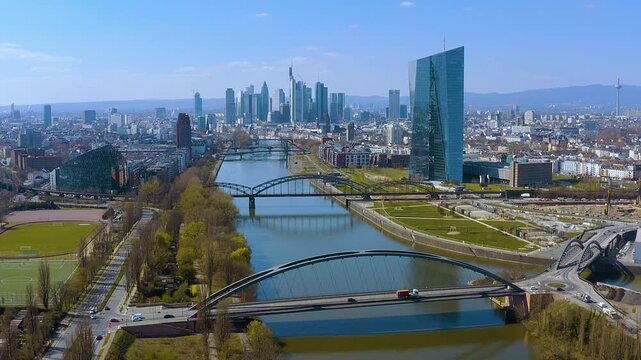 4K drone view of Frankfurt am Main with the European Central Bank, River Main, and multiple bridges leading to the modern skyline under a bright blue sky, showcasing Germany&rsquo;s financial capital.