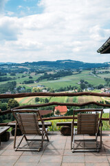 Two wooden lounge chairs on a terrace overlook a lush valley of fields, trees, and distant homes....