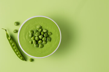 Green pea puree soup in a white bowl decorated with peas stands on a green background next to the...