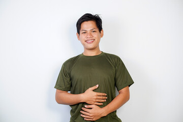 Obraz premium A cheerful young Asian man wearing a green shirt smiles while holding his stomach, standing against a plain white background, exuding happiness and good health. 