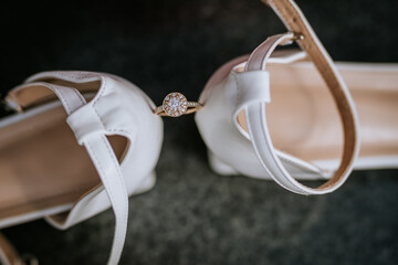 Elegant diamond engagement ring placed between white bridal shoes on a dark background, symbolizing...