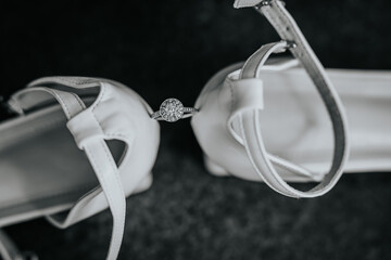 Black and white close-up of a diamond engagement ring placed between elegant white bridal shoes,...