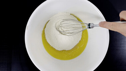 Flour added to melted butter in white mixing bowl with whisk, hand holding utensil on black table...