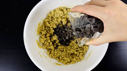 Adding chocolate chips to cookie dough in white bowl, hand pouring from glass over mixture on black...
