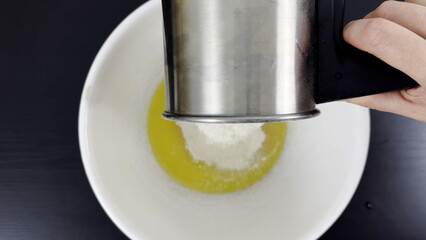 Flour being sifted into melted butter in white mixing bowl, hand holding stainless steel sifter...