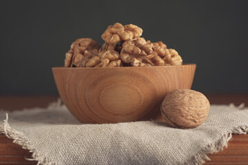 Shelled walnuts in a wooden bowl on a linen napkin and wooden table against a dark green background.