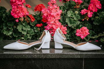 Elegant white bridal shoes with a diamond engagement ring placed between them, displayed against...
