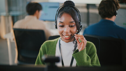 Confident African American female call center employee in headset offering customer support, tech consulting, CRM services, telemarketing, working at computer desk in diverse office workplace