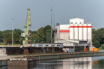 Industrial port with cranes and silos. A port area with a green gantry crane, white silos, and...