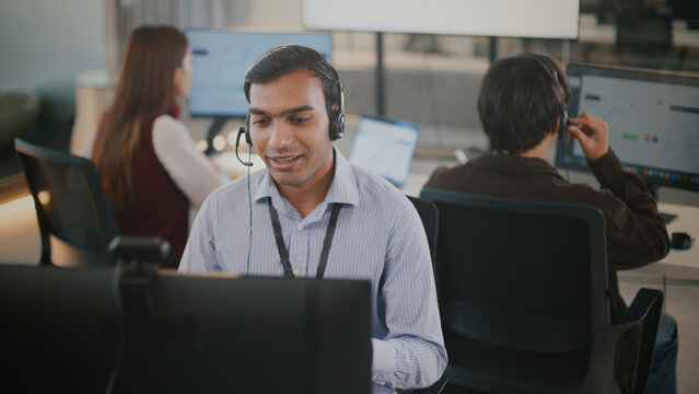 Confident Indian businessman provides customer service and tech support in a diverse corporate office. Wearing a headset, he speaks professionally with clients about CRM and business solutions.