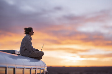 A woman works on her laptop atop a bus, set against a vibrant sunset. Symbolizing freedom, remote work, and digital nomadism. Ideal for travel, lifestyle, and tech themes.