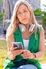 Young woman with headphones around her neck and smartphone in her hands, sitting on the grass in an...