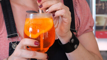 Woman sipping bright orange aperol spritz, garnished with citrus slice and ice cubes, relaxing...
