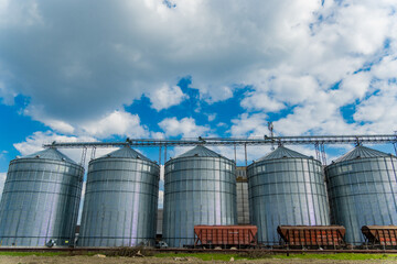 Tall metal silos stand in rows against a bright blue sky dotted with fluffy clouds, highlighting a farming location during midday