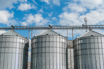 Three towering silos made of metal reflect sunlight, surrounded by a blue sky with scattered...