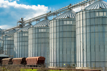 Several large silos are lined up neatly at a farming operation, with train cars parked nearby in...