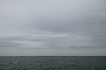Dramatic overcast sky stretches above a vast, calm sea. Wind turbines line the distant horizon,...