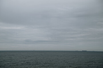 Dramatic overcast sky stretches above a vast, calm sea. Wind turbines line the distant horizon,...