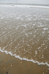 Waves crash on the sandy beach, under a moody sky. A red marker buoy and distant wind turbines mark the horizon over the North Sea.