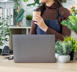 Remote Work. Young woman sipping coffee while standing at her desk in a plant-filled workspace.