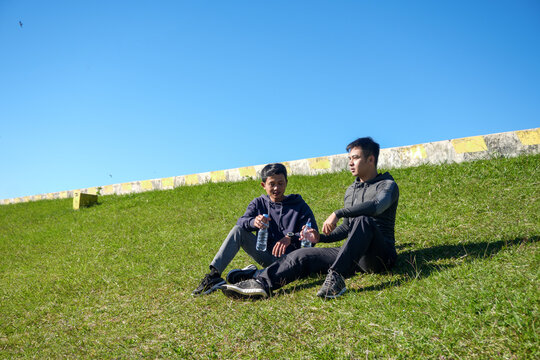 Two Young Men In Sportswear Rest On A Sunny Green Hill, Hydrating With Water After A Workout,