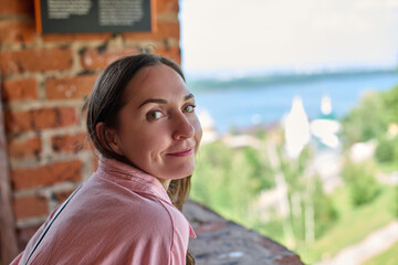 Young woman with long dark hair and pink shirt gazes over lush landscape from brick terrace....