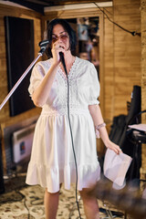 Young woman with black hair sings passionately in rustic studio. Warm lighting highlights wooden walls and cozy ambiance. Microphone and paper held confidently