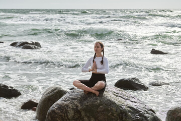 Young white woman with braids sits cross-legged on rocky shore. Overcast day with waves crashing, evoking tranquility and focus. Serene ocean backdrop enhances meditation scene
