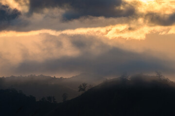 Morning light breaking through misty mountain forest ridge. Layers of mountains covered in early morning mist under a dramatic sky with warm sunlight breaking through clouds