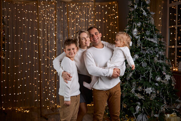 Happy young family parents and children gathering together on Christmas. Cheerful family of four in white sweaters smiles for a photo in a cozy, festive room and sparkling golden lights
