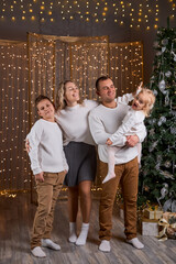 Happy young family parents and children gathering together on Christmas. Cheerful family of four in white sweaters smiles for a photo in a cozy, festive room and sparkling golden lights