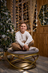 A smiling boy in a white sweater sits thoughtfully for a holiday photo, with a decorated Christmas...