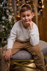 A smiling boy in a white sweater sits thoughtfully for a holiday photo, with a decorated Christmas...